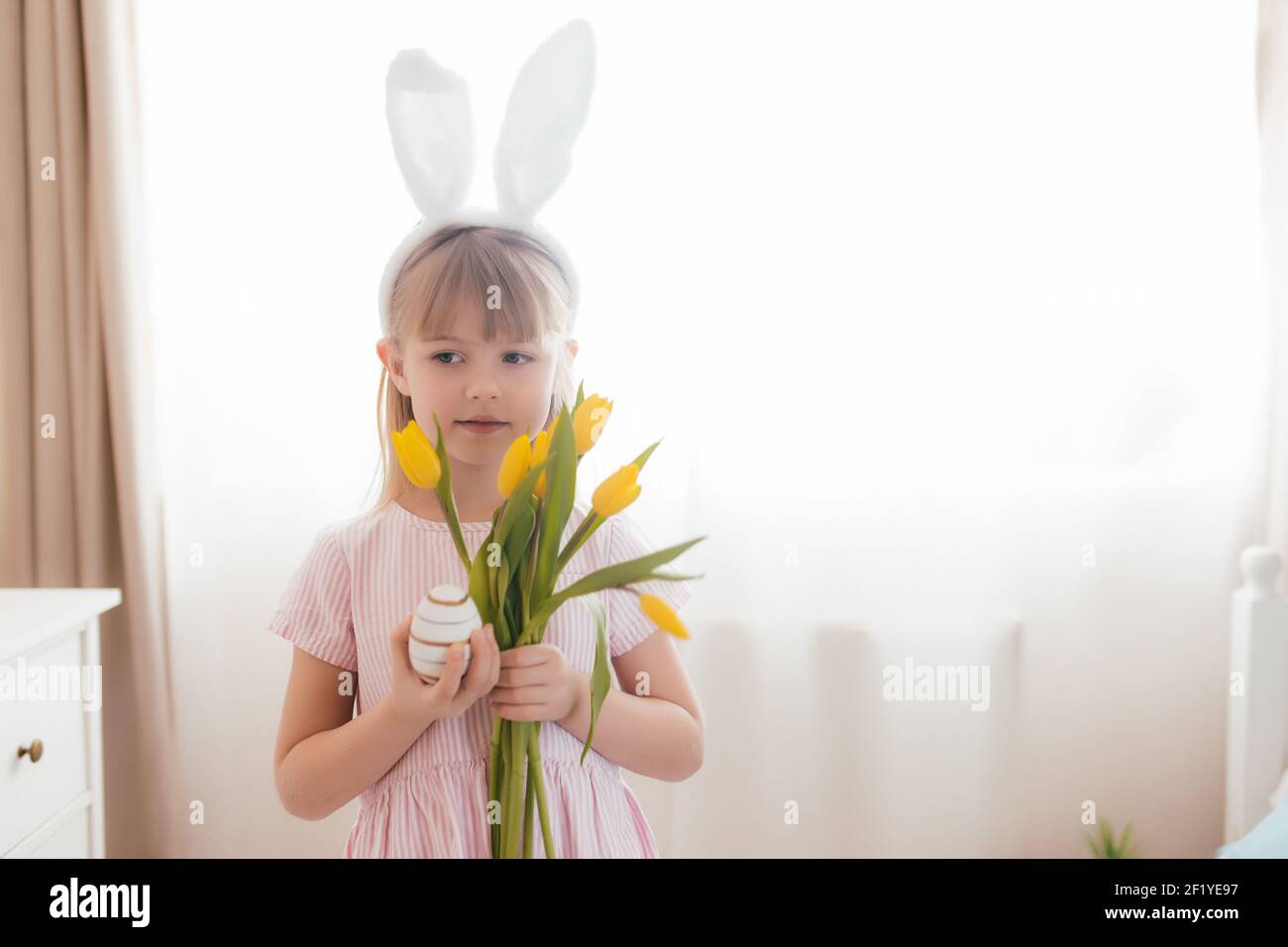 Osterkonzept. Kleines lächelndes Mädchen in weißen flauschigen Häschen Ohren und rosa Kleid mit gelben Tulpen Blumen und bunten Ei. Zimmereinrichtung. Hohe Qualität Stockfoto