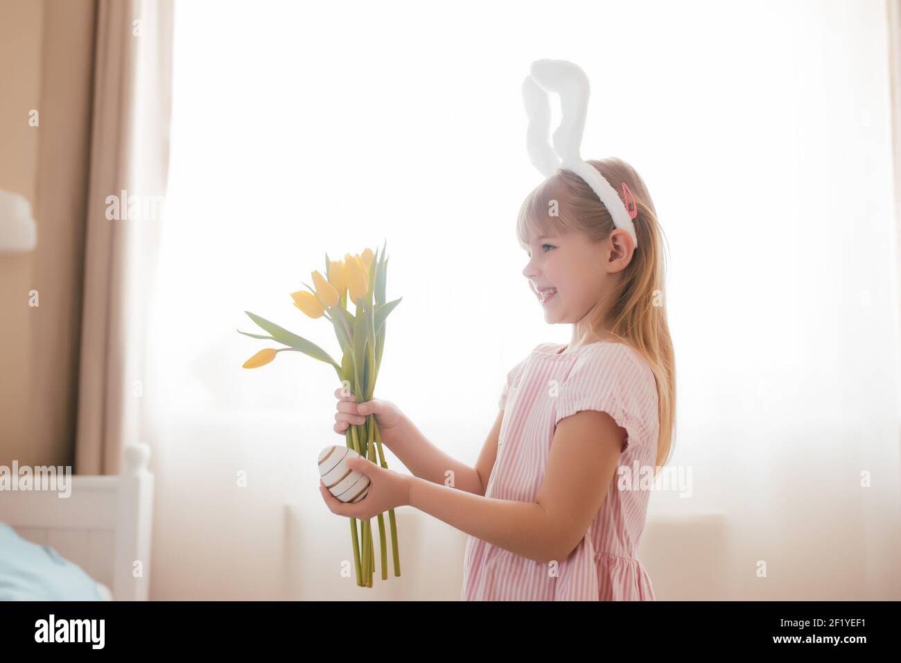 Osterkonzept. Kleines lächelndes Mädchen in weißen flauschigen Häschen Ohren und rosa Kleid mit gelben Tulpen Blumen und bunten Ei. Zimmereinrichtung. Hohe Qualität Stockfoto