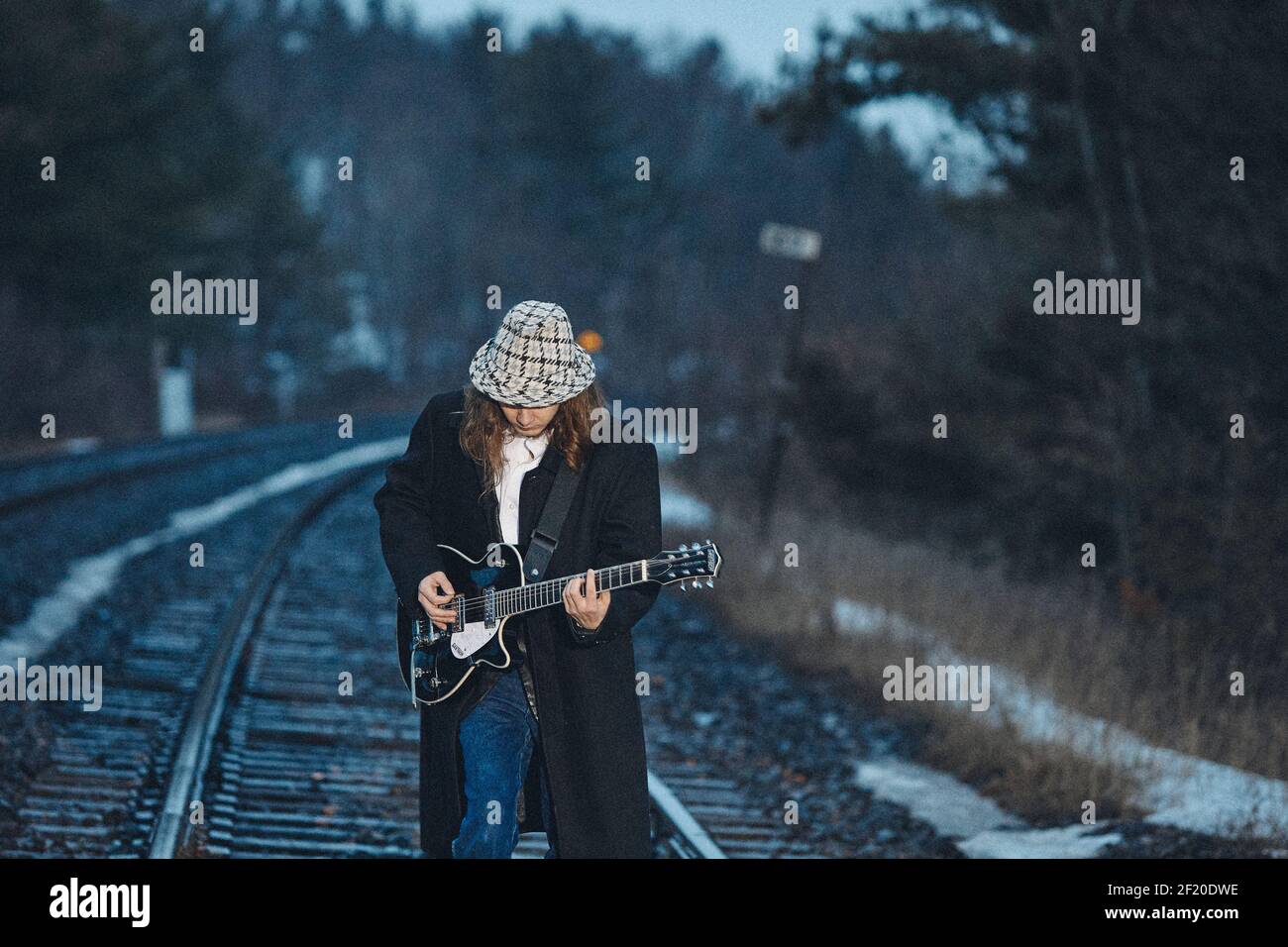 Junger Mann in Hut und Mantel, der auf Schienen steht und Spielen E-Gitarre am Abend auf dem Land Stockfoto