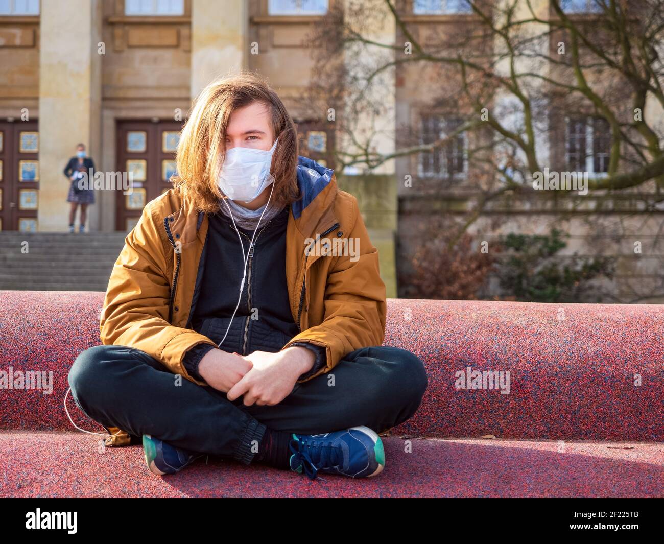 Trauriger junger Mann trägt schützende medizinische Maske, um sich zu schützen Von Pandemie Coronavirus im Freien und sitzen allein in einer Stadt Und Musik hören Stockfoto