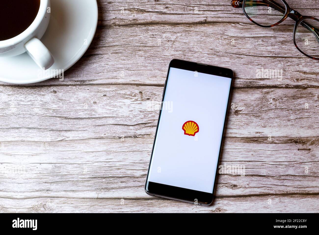 Ein Handy oder Handy auf einem Holz gelegt Tisch mit der Shell-App neben einem Kaffee geöffnet Und Brille Stockfoto