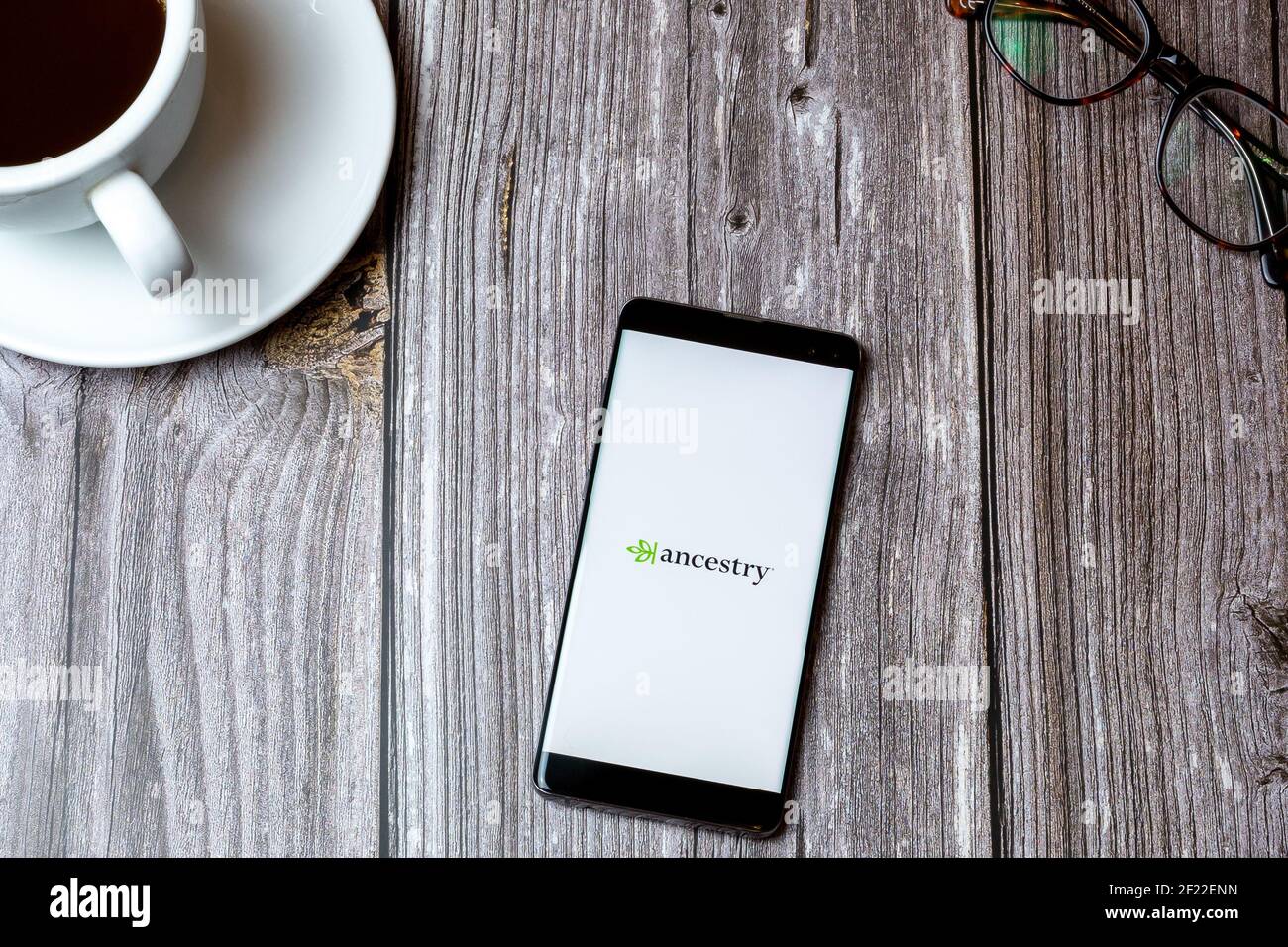 Ein Handy oder Handy auf einem Holz gelegt Tabelle mit der App Ancestry auf dem Bildschirm neben geöffnet Ein Kaffee Stockfoto