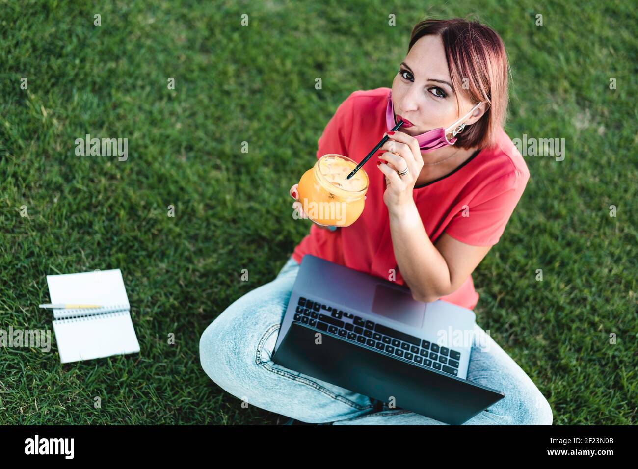 Frau 40 Jahre alt arbeitet im Freien mit Laptop während Coronavirus Outbreak - lächelnde Unternehmerin sitzt auf Rasen mit pc Während Sie Saft trinken Stockfoto