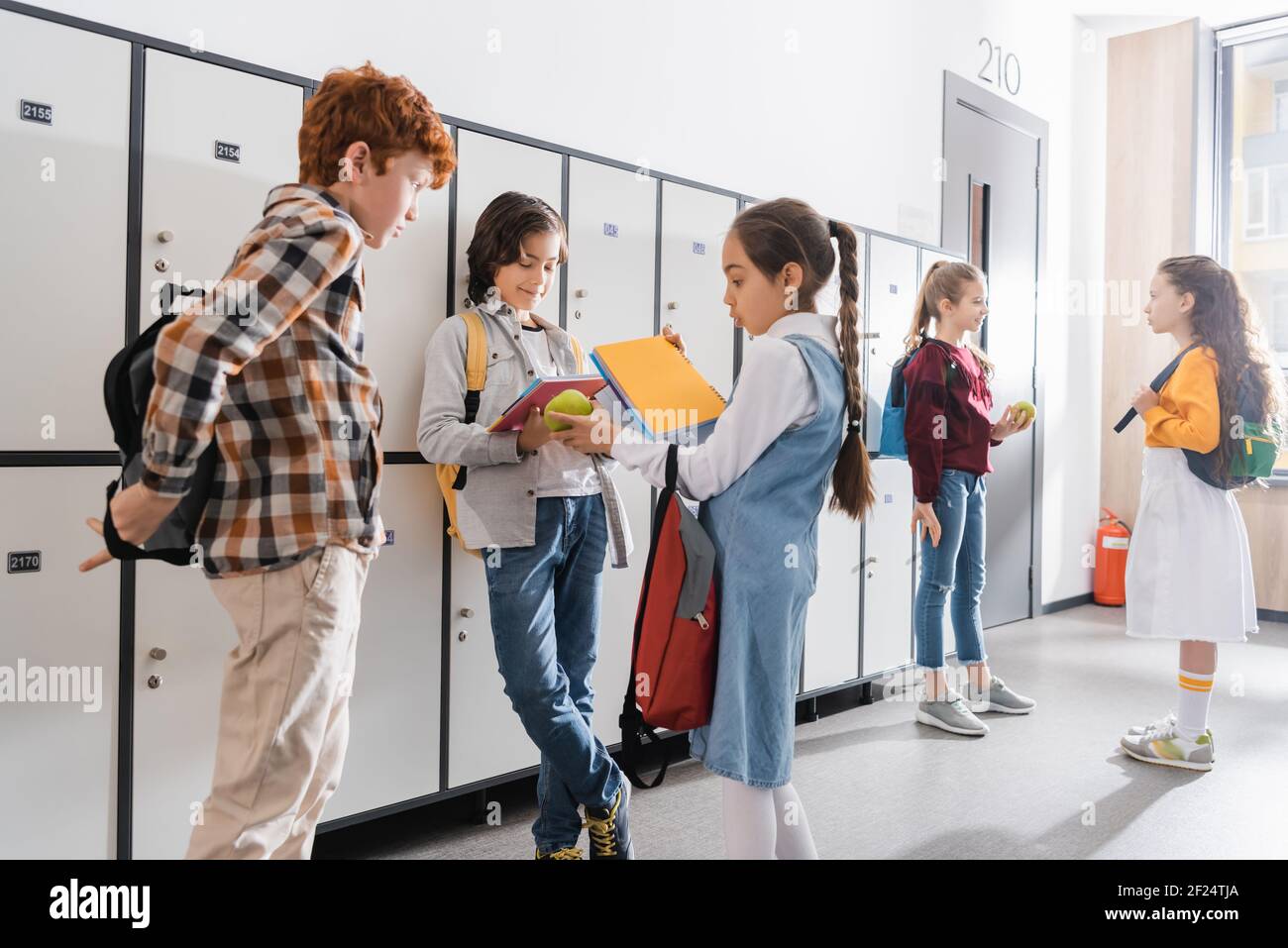 Schulmädchen hält Apfel in der Nähe Jungen mit Rucksäcken in der Schule Stockfoto