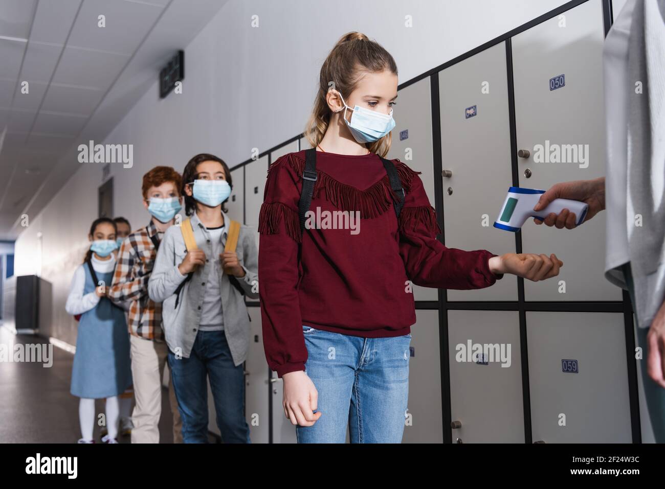 Lehrer Messung der Temperatur von Schulmädchen in medizinische Maske mit Infrarot Thermometer in der Halle Stockfoto