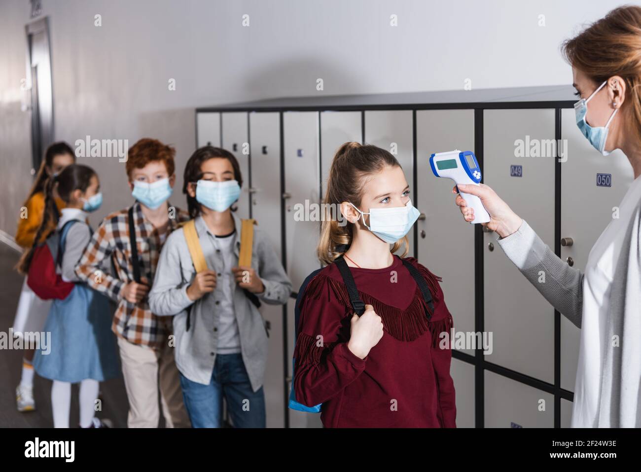Lehrer mit Infrarot-Thermometer bei der Überprüfung der Temperatur des Schulkindes in Medizinische Maske in der Halle Stockfoto
