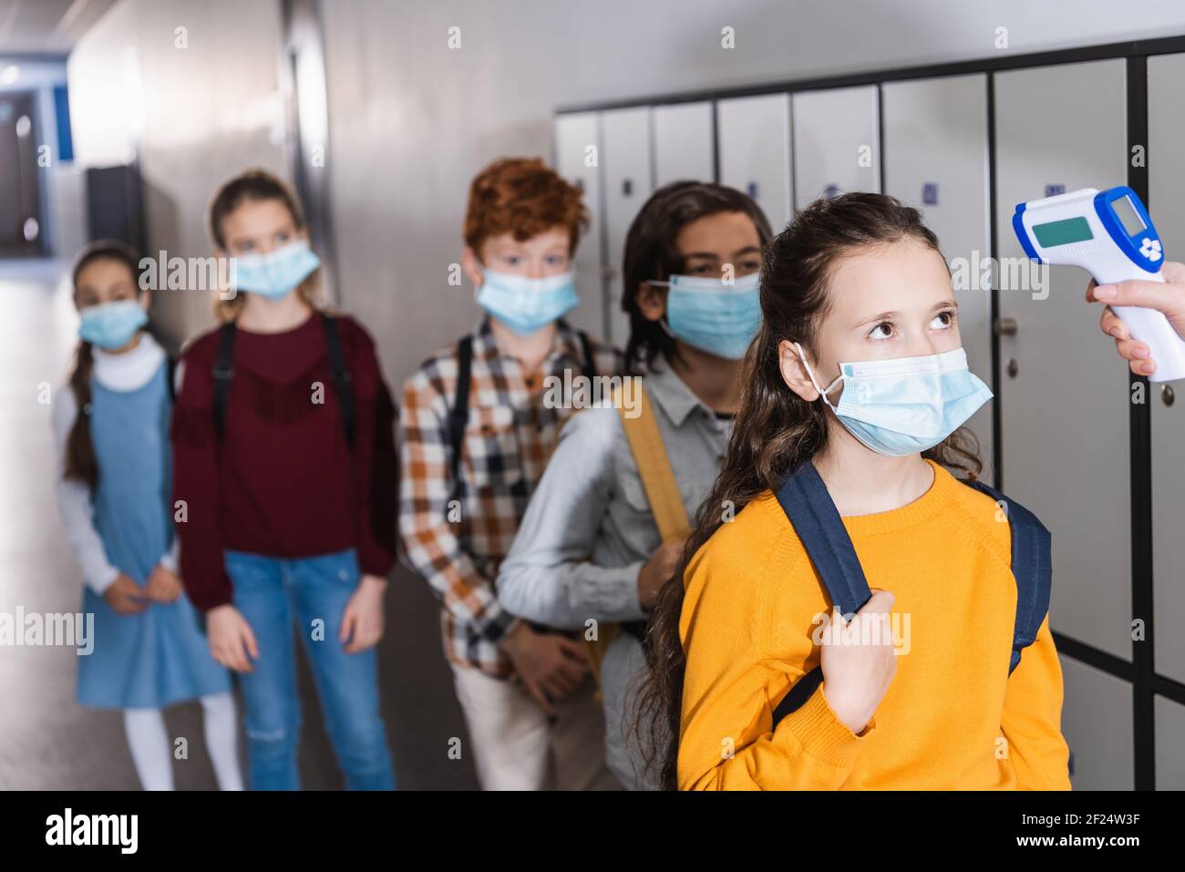 Lehrer mit Infrarot-Thermometer auf Schüler in der medizinischen Maske in der Nähe Kinder auf unscharfem Hintergrund Stockfoto