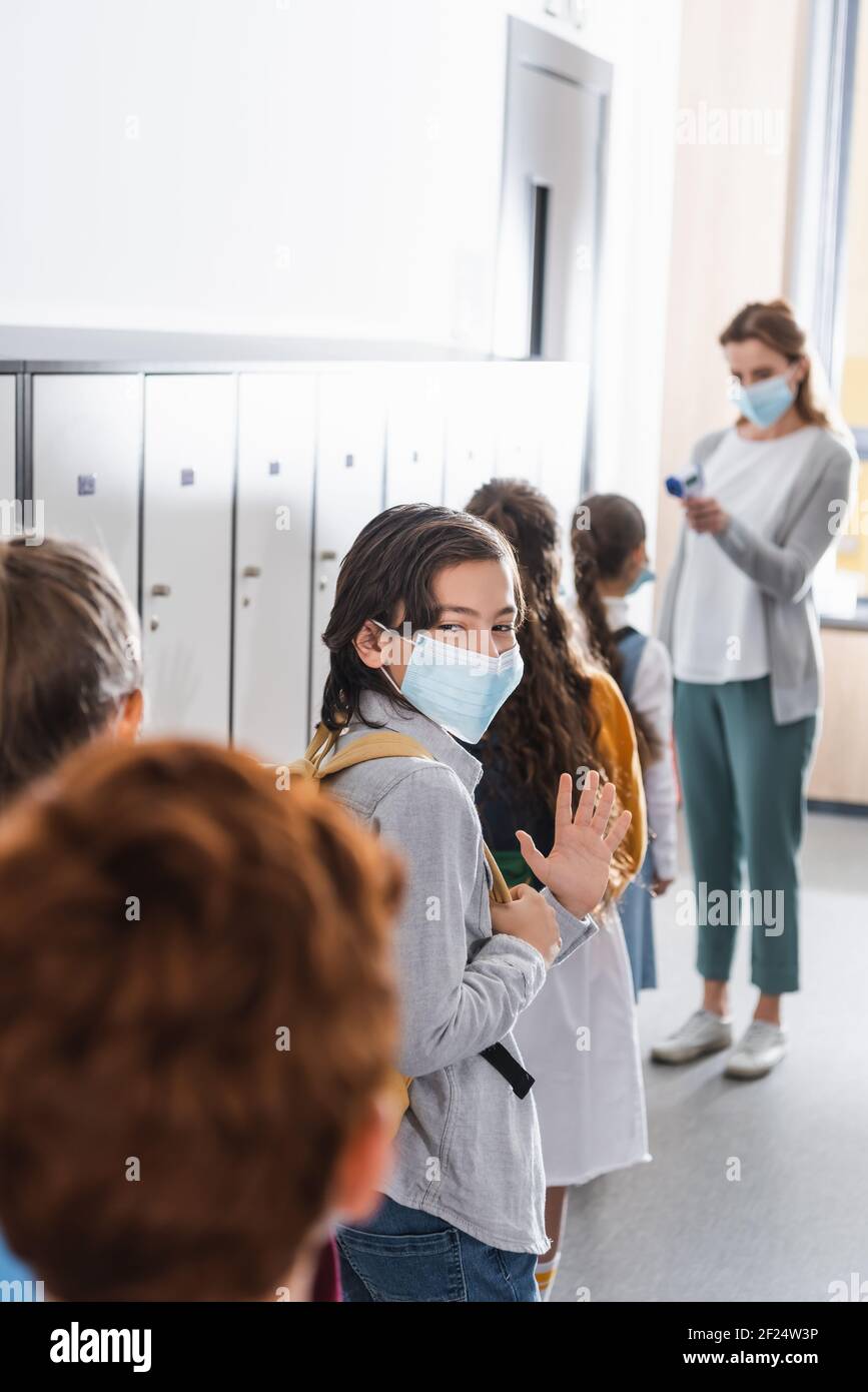 Junge in medizinischer Maske winkende Hand in der Nähe von Klassenkameraden und Lehrer Mit Infrarot-Thermometer auf verschwommenem Hintergrund Stockfoto