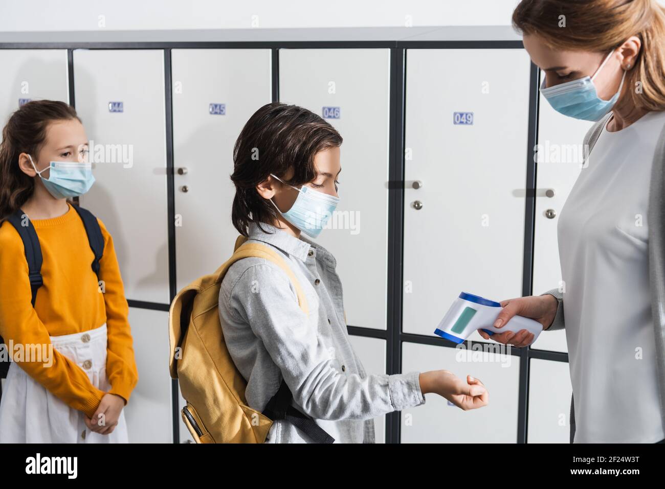 Lehrer in der medizinischen Maske mit Infrarot-Thermometer auf der Hand Pupille im Korridor Stockfoto