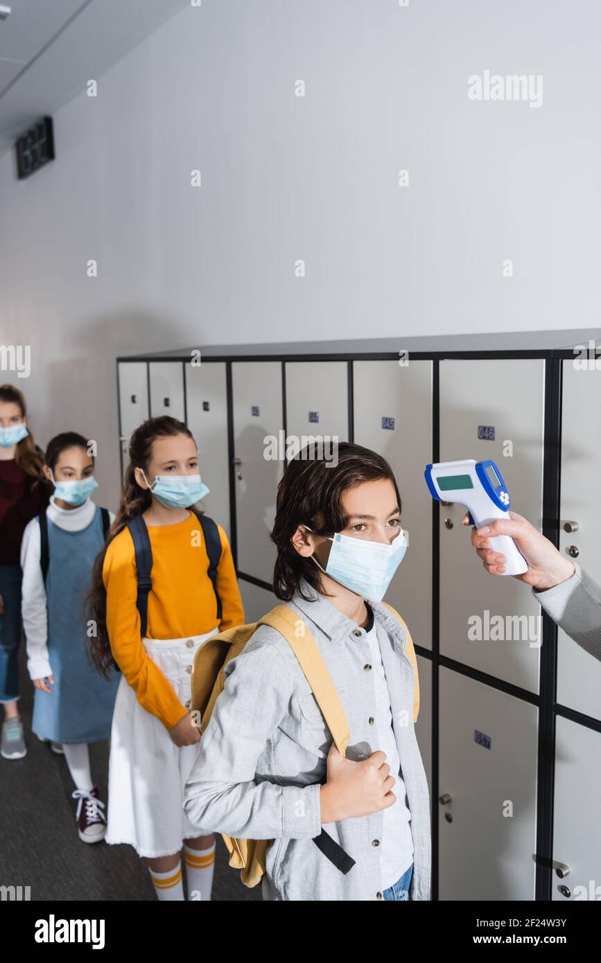 Lehrer mit Infrarot-Thermometer auf Schuljunge in Schutzmaske in der Nähe Kinder auf unscharfem Hintergrund Stockfoto