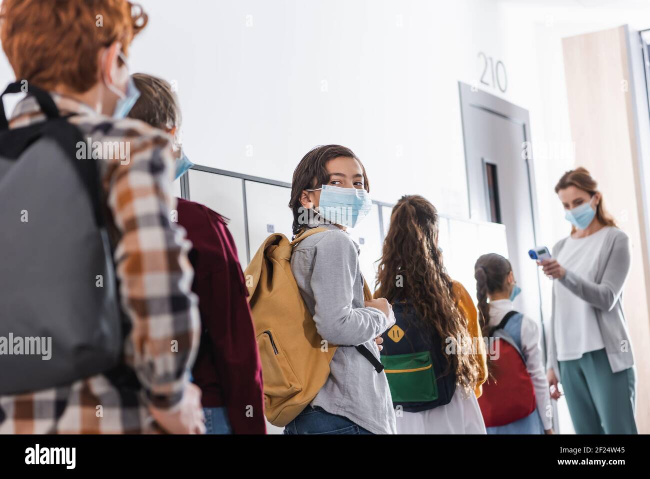 Junge in medizinische Maske in der Warteschlange in der Nähe von Klassenkameraden und stehen Lehrer mit Infrarot-Thermometer auf verschwommenem Hintergrund Stockfoto
