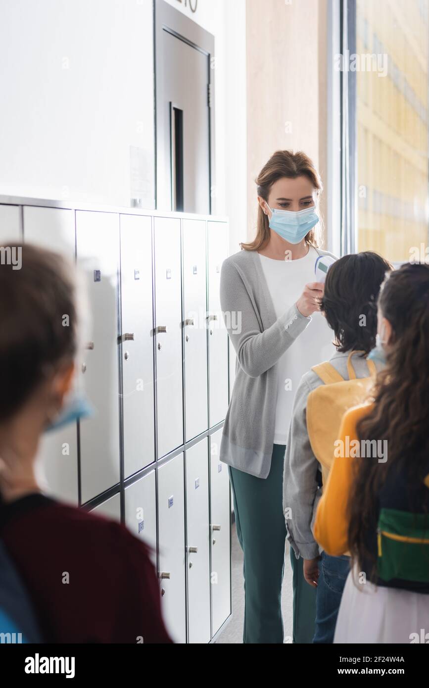 Lehrer in der medizinischen Maske, die die Temperatur der Schüler in der Nähe der Schließfächer misst In der Schule Stockfoto