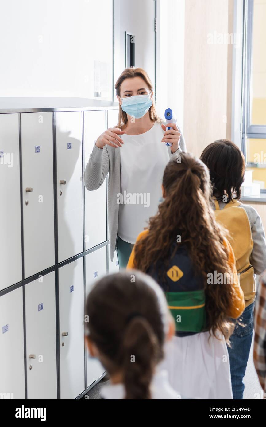 Lehrer in der medizinischen Maske hält Infrarot-Thermometer in der Nähe der Schüler in Warteschlange im unscharfen Vordergrund Stockfoto