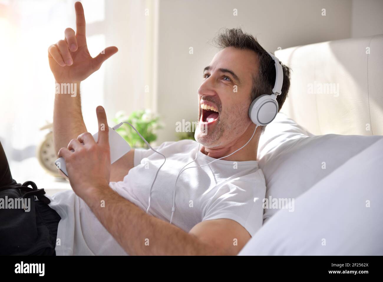 Euphorischer Erwachsener, der im Bett liegt und Musik hört Von einem Handy mit weißen Kopfhörern gekleidet in Schlafanzug mit Morgensonne kommt durch die Stockfoto