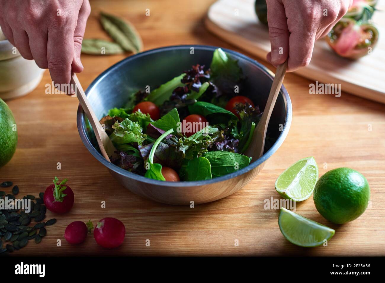 Menschenhände rühren einen Salat in eine Metallschüssel, mit etwas Holzbesteck, auf einem Holztisch in einer Küche. In der gastronomischen Fotografie können Sie radis sehen Stockfoto