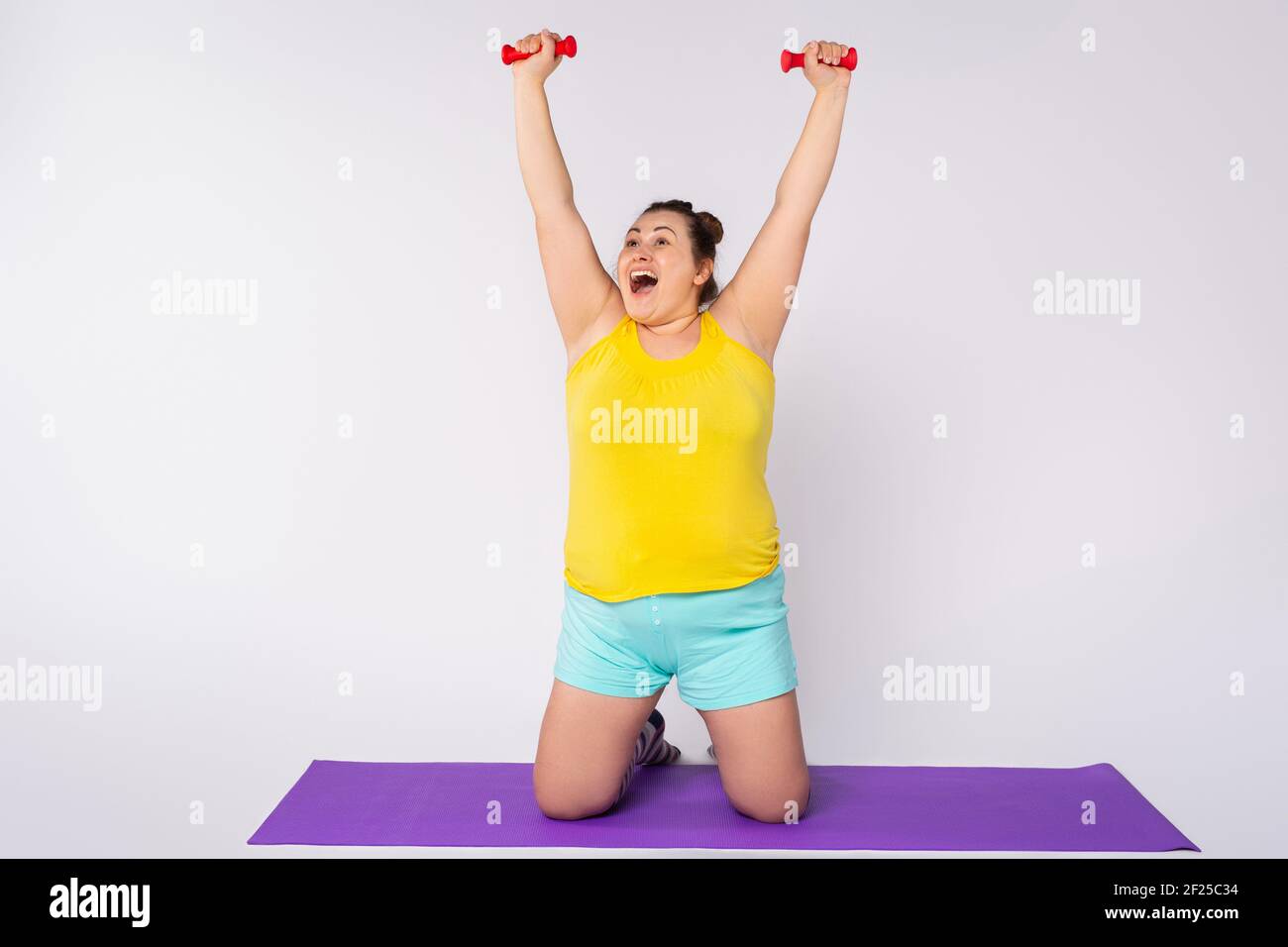 Glückliche Plus Größe Frau in hellen Hauskleidung tun Übungen mit blauen Hanteln auf den Knien auf einer Yoga-Matte. Isoliert auf grauem Hintergrund mit Seite Stockfoto