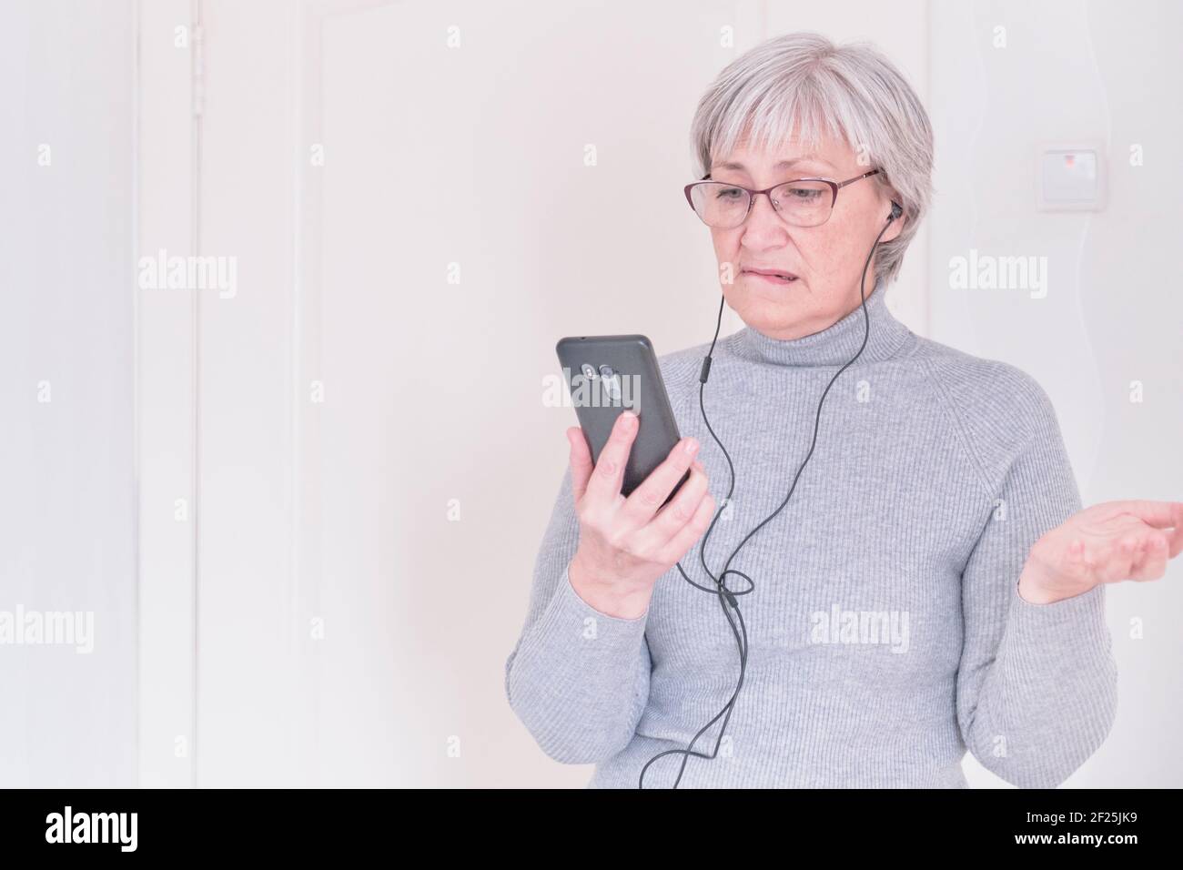 Eine grauhaarige ältere Frau mit Brille und einem grauen Rollkragen, die zu Hause mit dem Handy nachdenklich auf die Lippe beißt. Menschen verbinden, communicatio Stockfoto
