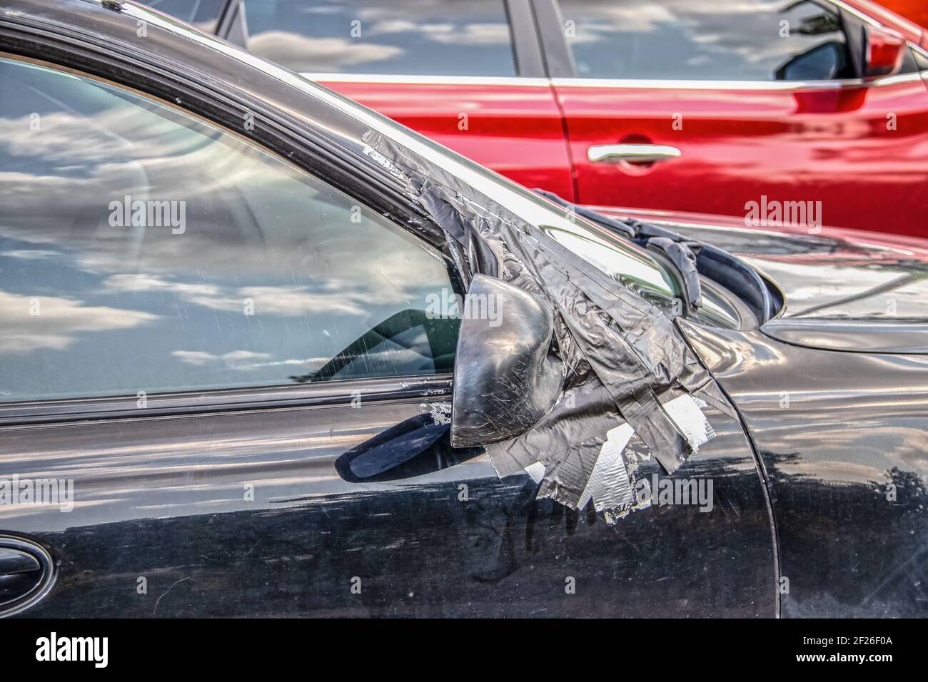 Nahaufnahme eines schmutzigen Autos mit aufgeklebtem rechten Seitenspiegel Und Kratzer an ihm und Fenster - viele Reflexionen Von blauem Himmel und einem leuchtend roten verschwommenen Auto in Stockfoto