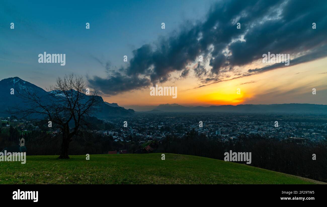 Sonnenuntergang mit Nachglühen über dem Rheintal. Dornbirn liegt im nebligen Tal, Schweizer Berge sind oben. Einzelner Baum steht auf der abfallenden Wiese Stockfoto