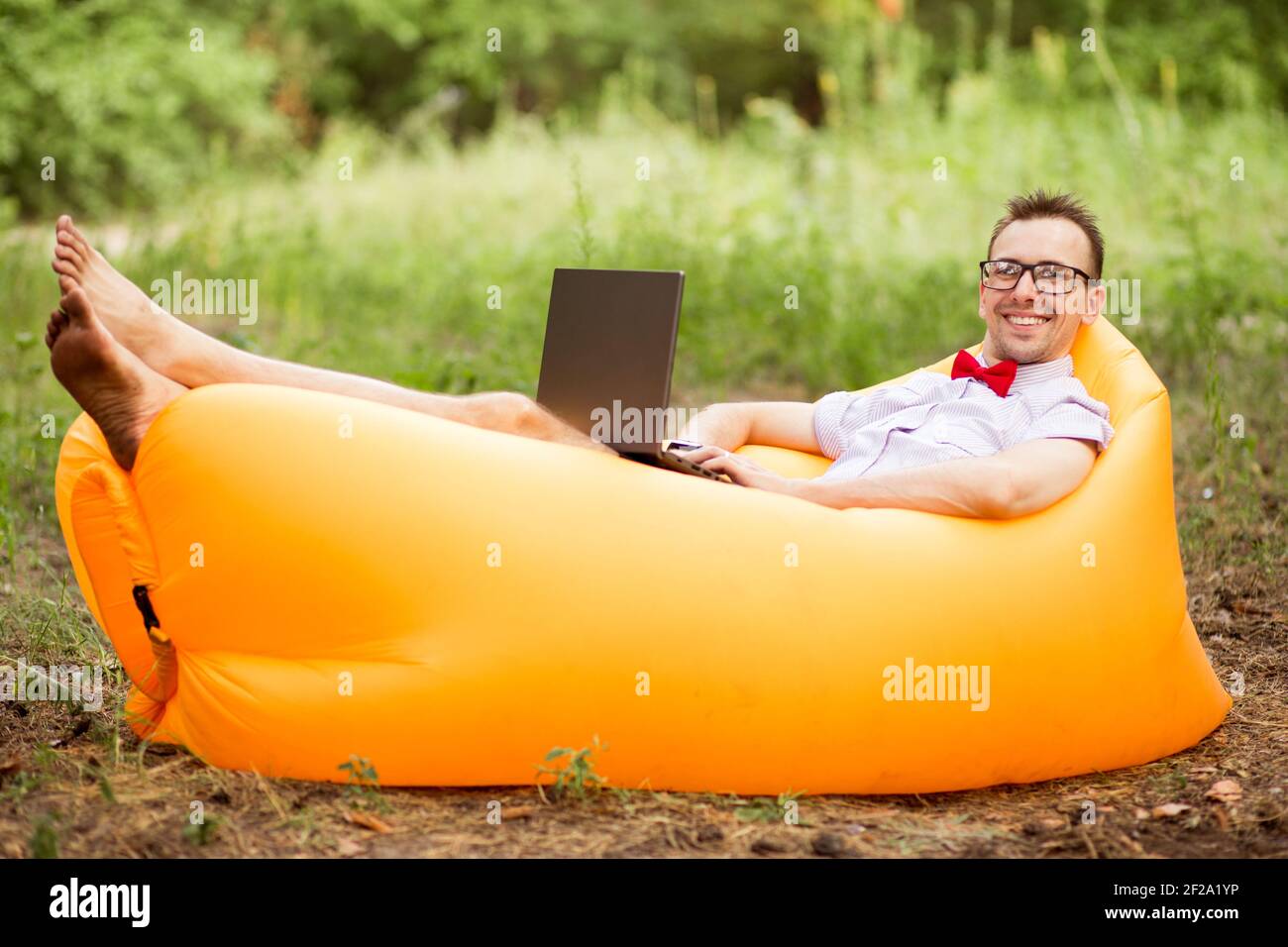 Glücklicher Mann in der Brille arbeitet mit Laptop im Sommerpark auf aufblasbarer Matratze. Konzept für freiberufliche und Telearbeit Stockfoto