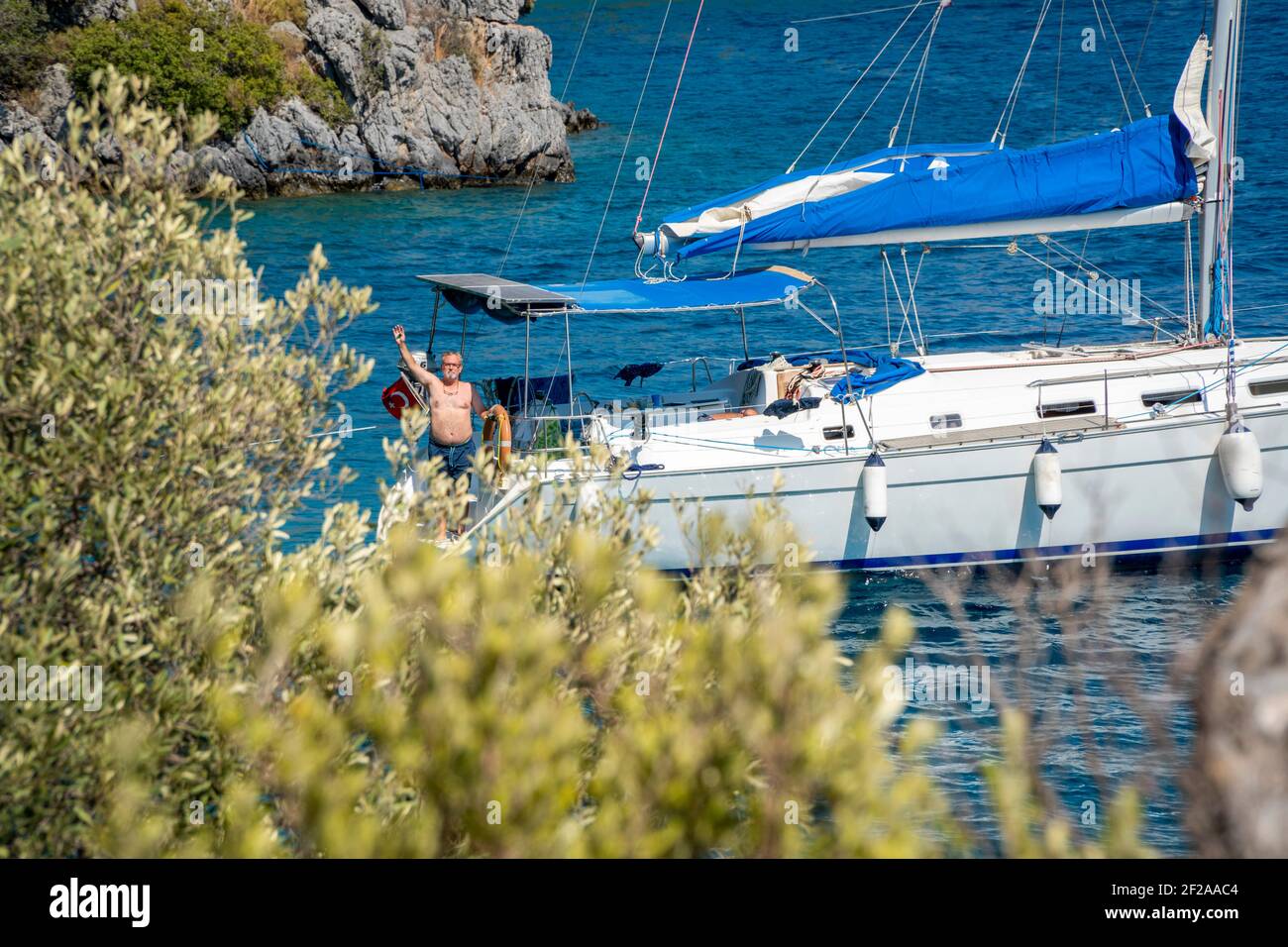 Hinter einem grünen Baum ein einzelnes Segelschiff mit einem Mann. Die Person trägt Badeshorts und winkt der Kamera zu. Blaues Wasser der Ägäis Stockfoto