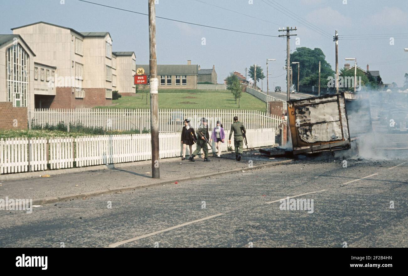 BELFAST, VEREINIGTES KÖNIGREICH - JUNI 1972. Schulkinder, die während der Unruhen vor Soldaten der britischen Armee vorbeikamen und ein brennendes entführtes Fahrzeug, Ballymurphy, West-Belfast, Nordirland, 1970er Jahre Stockfoto