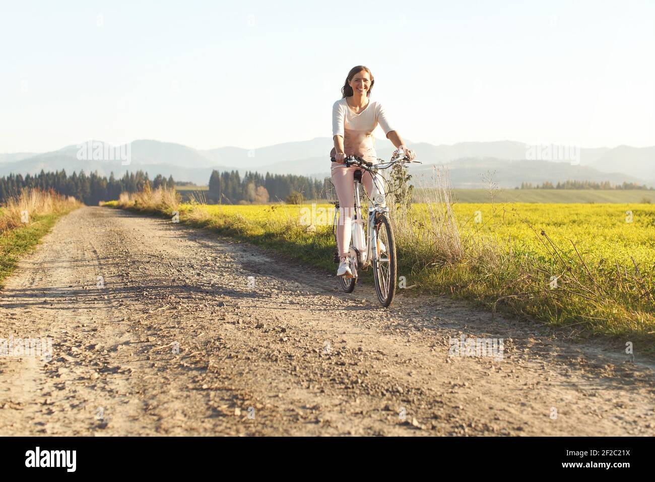 Junge Frau fährt Fahrrad auf staubiger Landstraße, lächelnd, Nachmittagssonne scheint auf sie, Blick von vorne Stockfoto