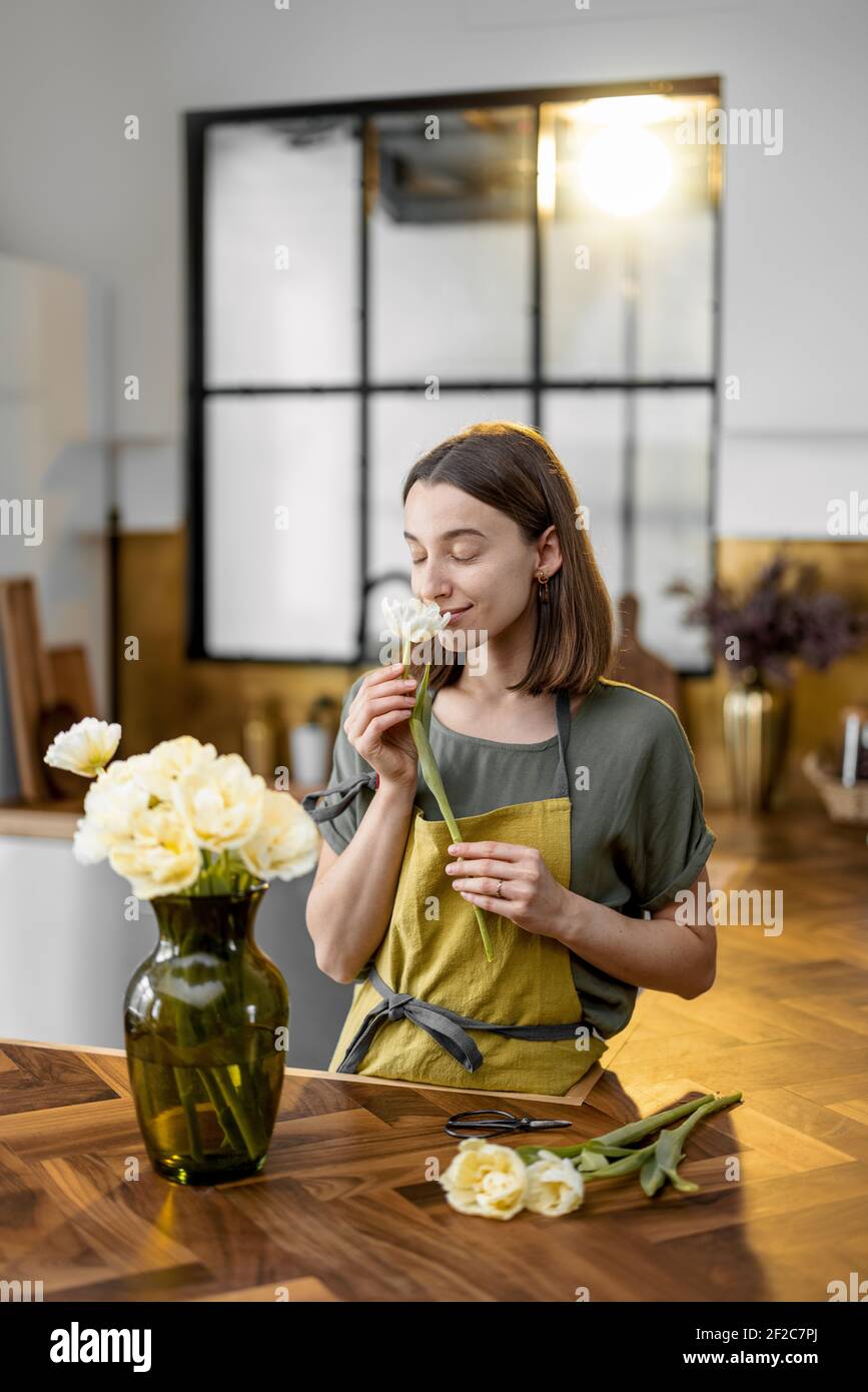 Die junge Frau atmet den Duft frischer Tulpen in grüner Schürze ein, während sie die Inneneinrichtung mit einem Bouquet frischer Blumen schmückt. Floristen mit Blumen auf der Küche zu Hause. Stockfoto