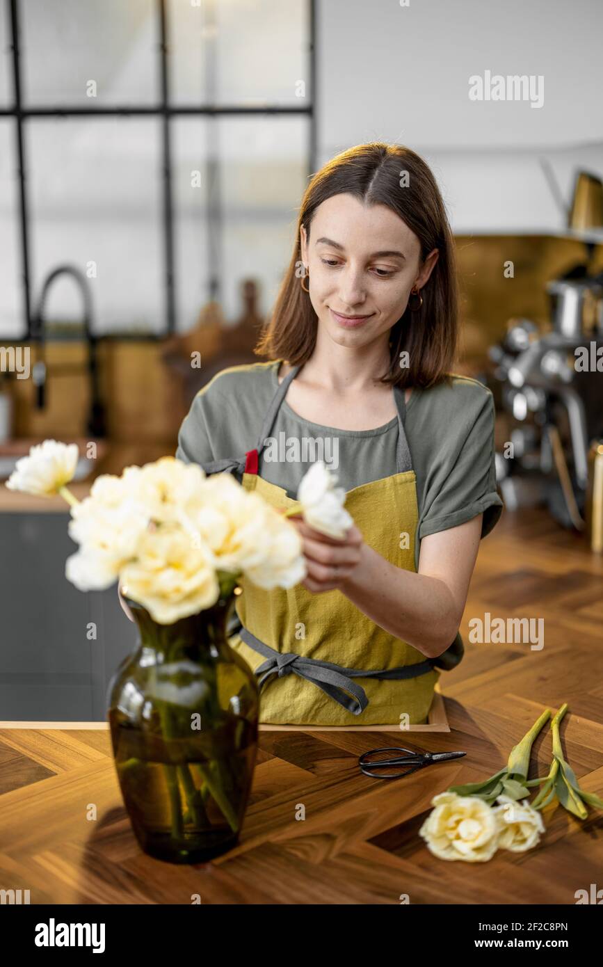 Die junge Hausfrau in grüner Schürze schmückt die Inneneinrichtung mit einem Bouquet frischer Tulpen. Floristen mit Blumen auf der Küche zu Hause. Setzen Blumen an Vase. Stockfoto