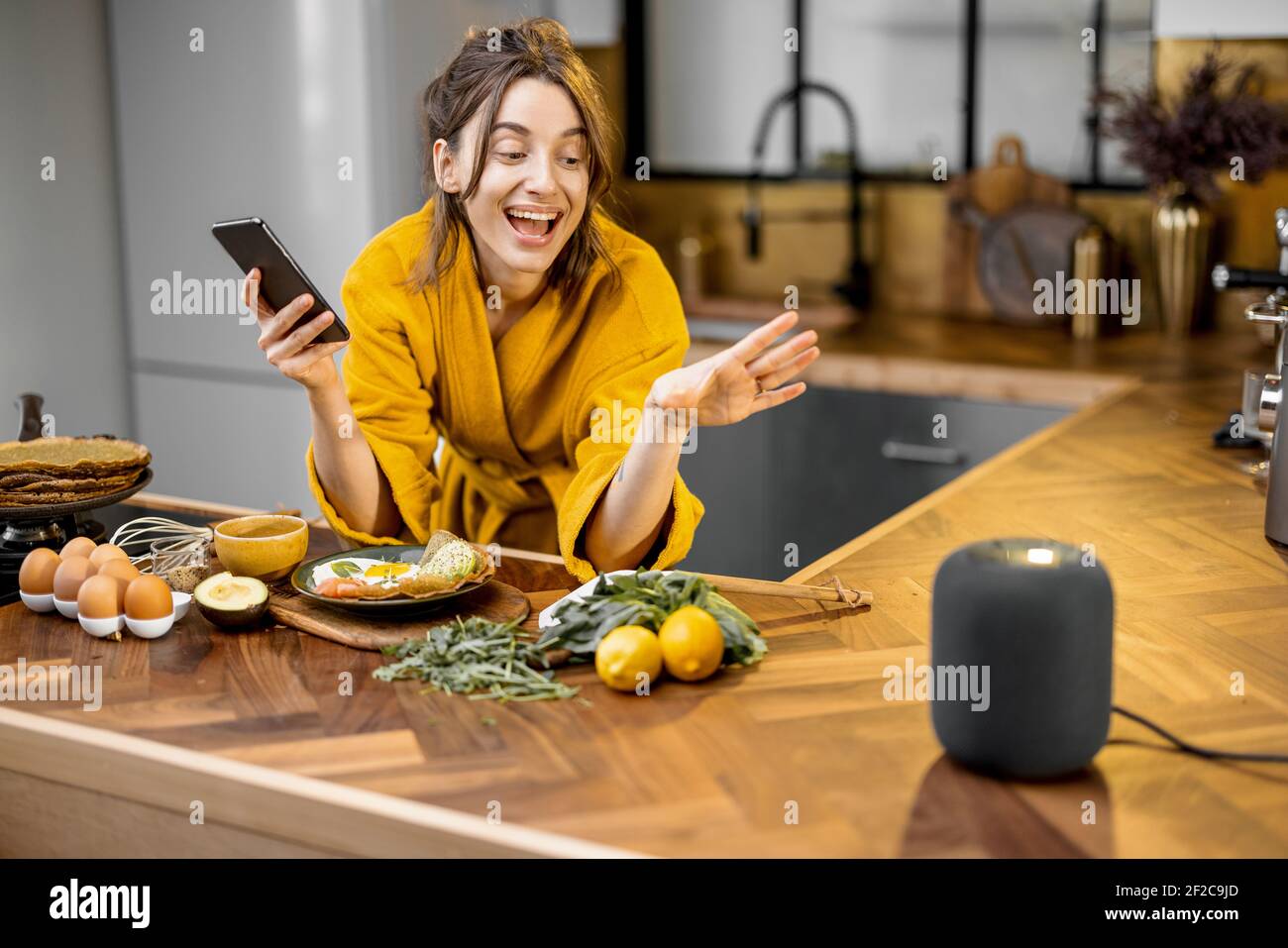 Glückliche Frau im Bademantel Steuerung Heimgeräte mit einer Sprachbefehle, sprechen zu einer intelligenten Spalte während der Frühstückszeit in der Küche zu Hause. Smart Home-Konzept Stockfoto