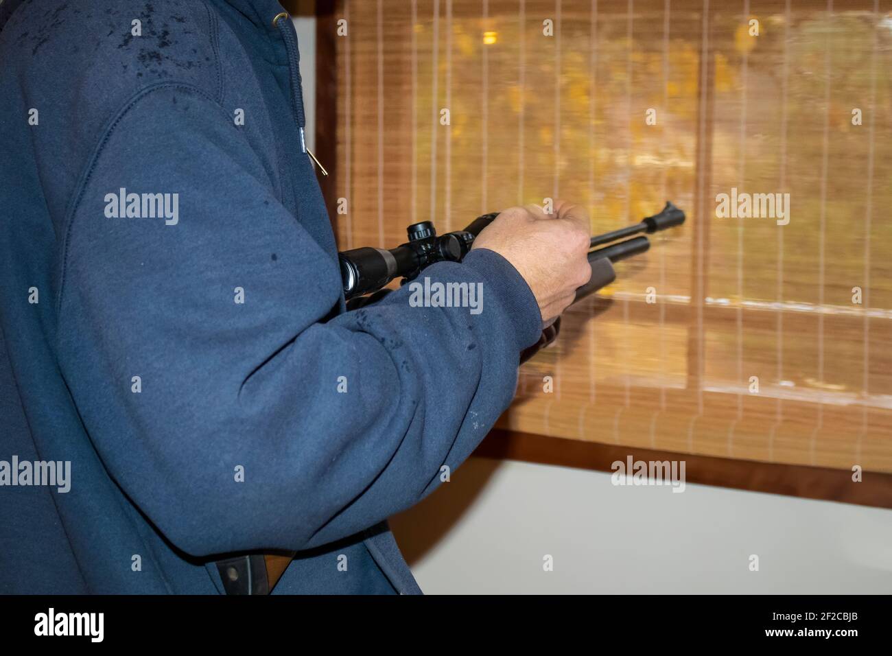 Kurzer Mann in Kapuzenjacke mit Luftgewehr im Stehen Durch ein Fenster mit dem Wald außen durch gewebt zeigen Schatten Stockfoto