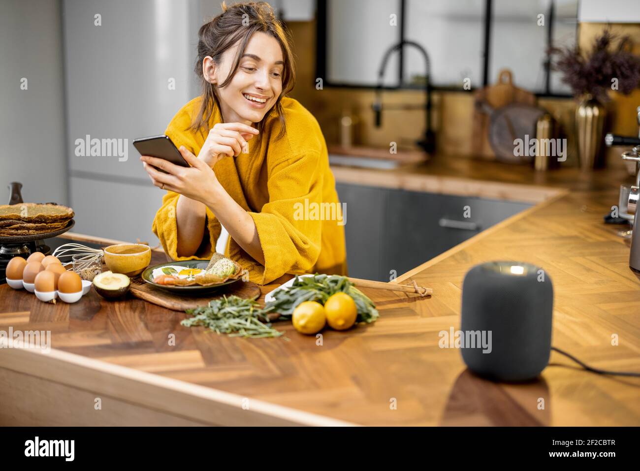 Glückliche Frau im Bademantel Steuerung Heimgeräte mit einer Sprachbefehle, sprechen zu einer intelligenten Spalte während der Frühstückszeit in der Küche zu Hause. Smart Home-Konzept Stockfoto