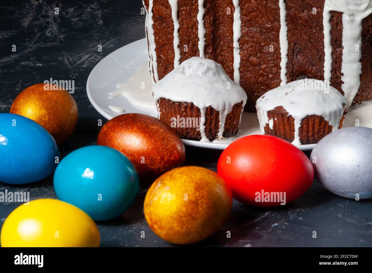 Hausgemachte handgemachte Osterkuchen mit weißer Glasur und bunt bemalten Ostereiern auf Tellern auf dunklem Hintergrund bedeckt. Stockfoto
