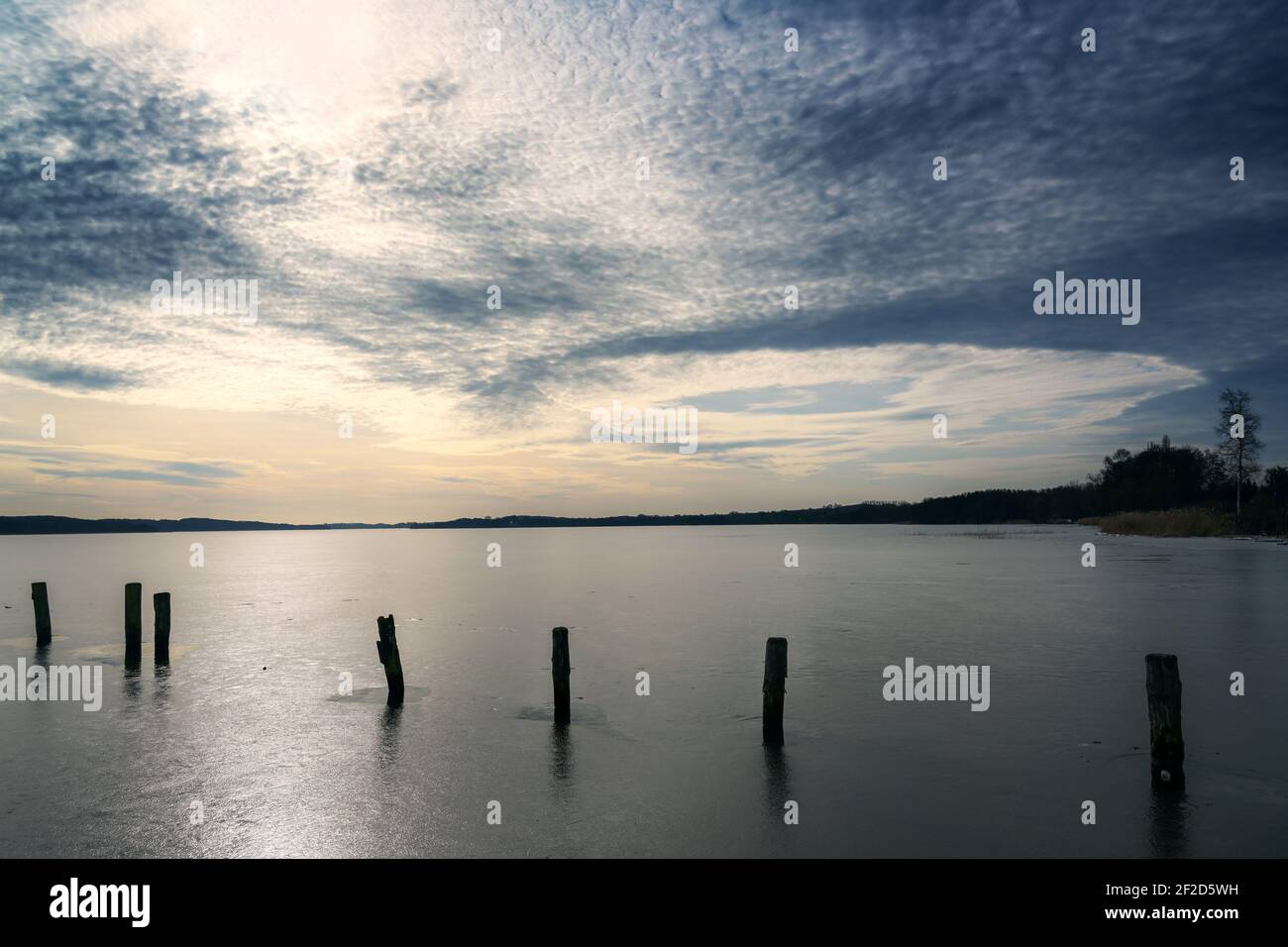 Die Reihe der dunklen hölzernen Pfähle auf dem gefrorenen See unter dem Himmel mit den interessanten Wolken, im späten Winter steht die Erde still, während es schon die Bewegung i gibt Stockfoto