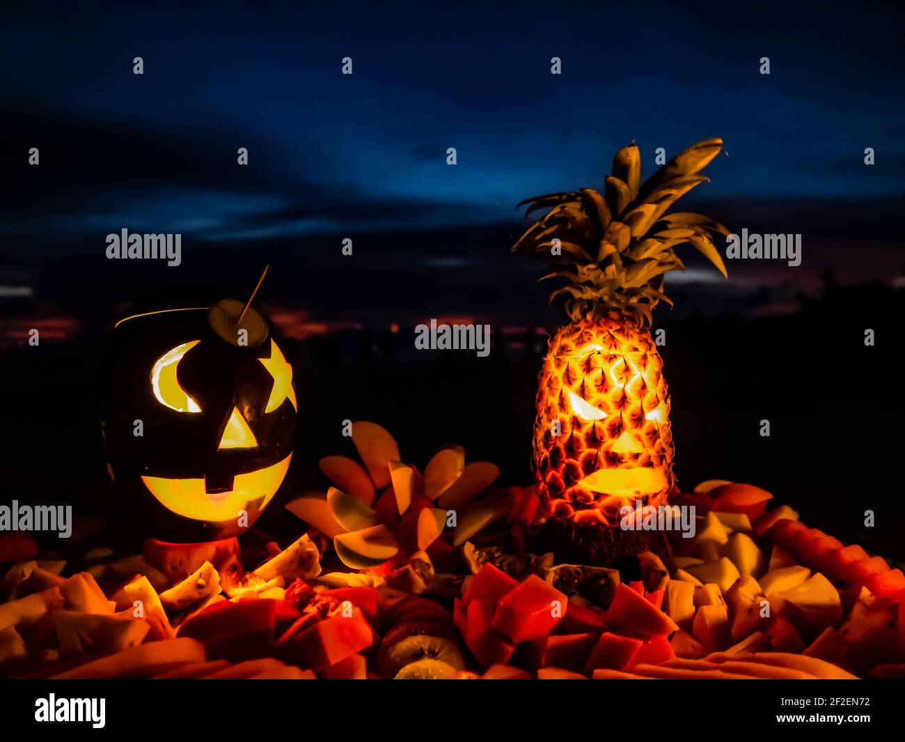 Silhouetten der Köpfe von fabelhaften Kreaturen aus Kürbis und Ananas geschnitzt, von innen durch Kerzen auf einem Tablett mit Obst gegen die Nacht beleuchtet Stockfoto