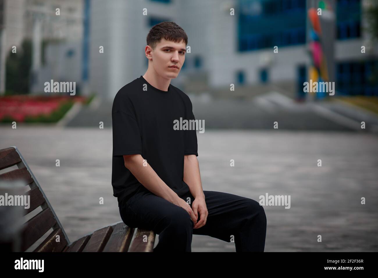 Schwarzes T-Shirt Mockup auf einem jungen Kerl auf einer Bank auf einem verschwommenen Hintergrund im Park sitzen, stilvolle Kleidung für Design-Präsentation. Leeres legeres Gerinnsel Stockfoto