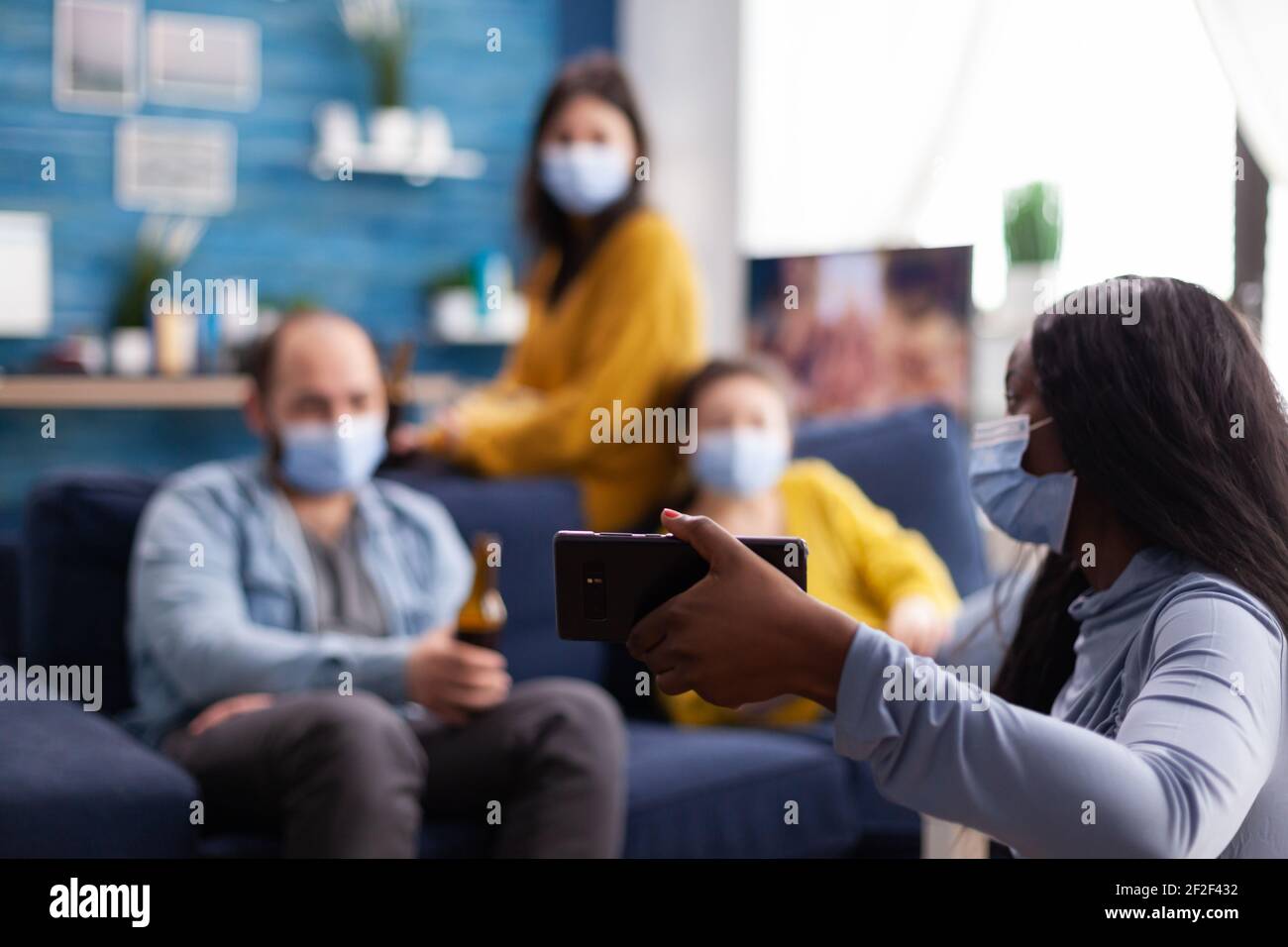 Afrikanische Frau zeigt Videoclip zu fröhlichen Freunden auf Smartphone im Wohnzimmer zu Hause tragen Gesichtsmaske als Prävention gegen die Ausbreitung und Infektion von covid19 beim Abhängen. Konzeptuelles Image. Stockfoto