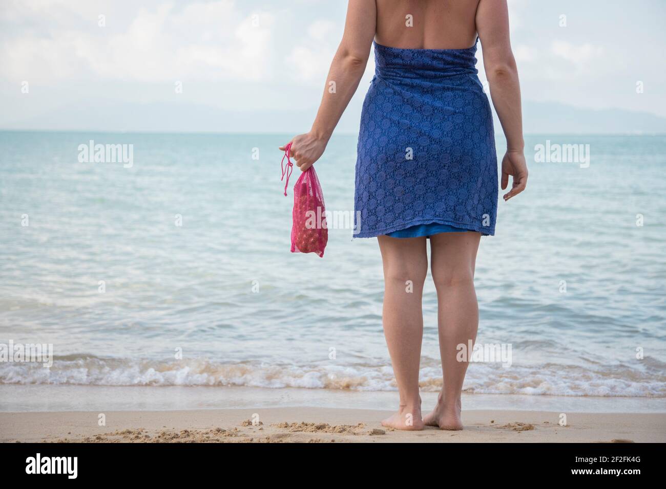 Eine Frau hält in der Hand eine Mesh-Einkaufstasche und steht am Rande des Meeres an einem Sandstrand. Ökologie-Konzept. Bewusster Konsum Stockfoto
