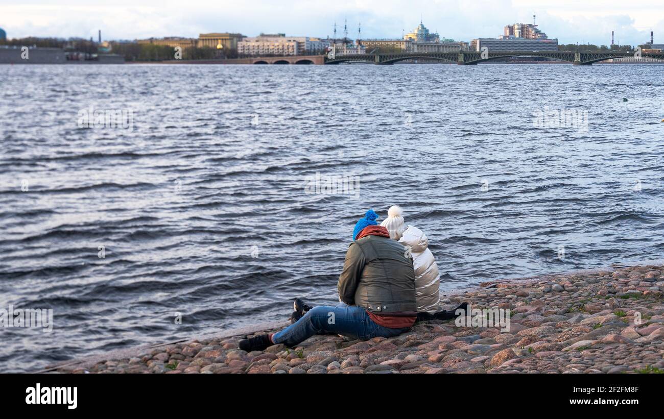 Zwei Personen in warmer Kleidung sitzen auf den Kopfsteinpflaster des Ufers des Flusses, Rückansicht Stockfoto