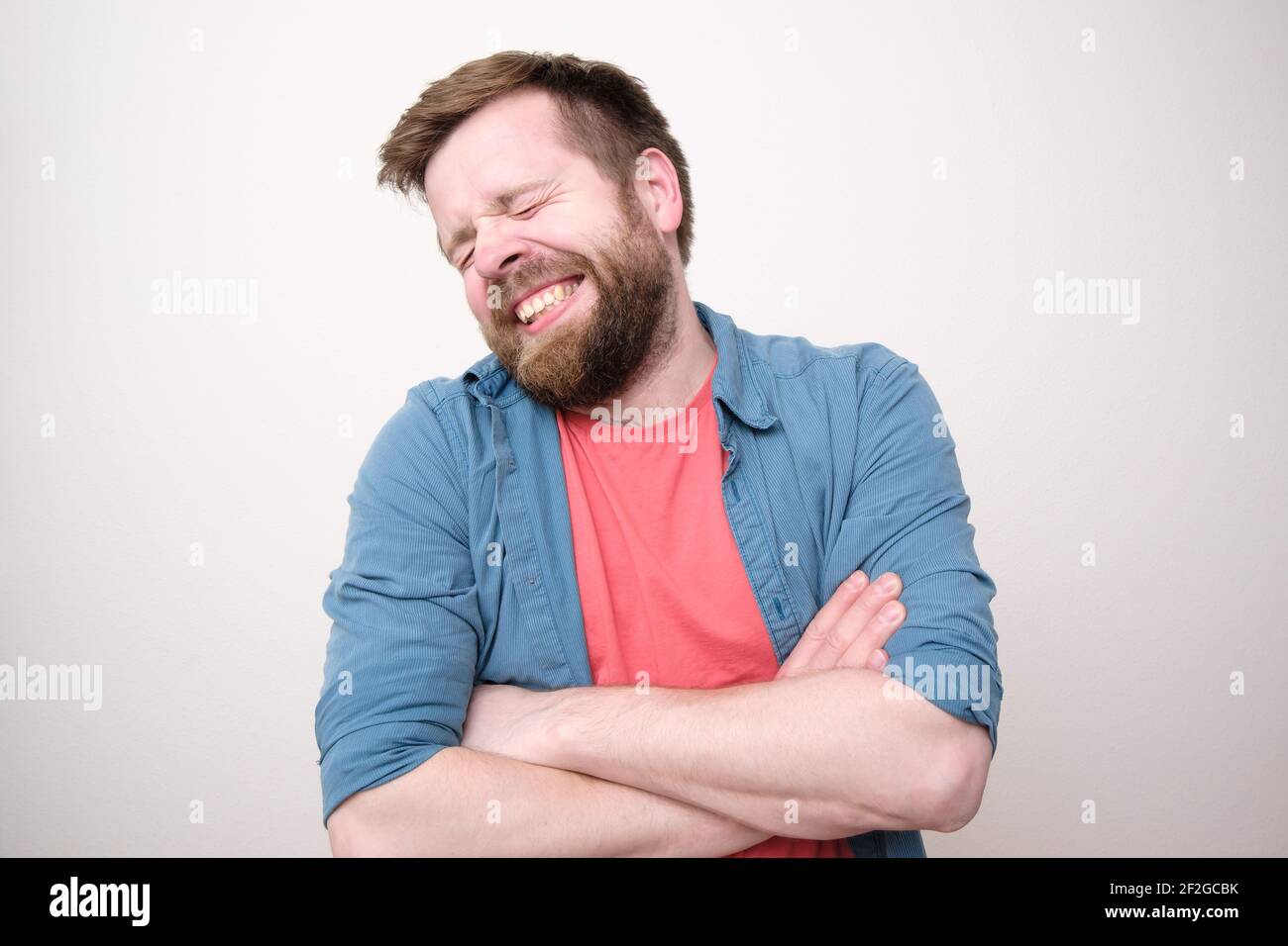Lustige schüchterne Mann steht mit gekreuzten Armen und lächelt niedlich mit geschlossenen Augen. Weißer Hintergrund. Stockfoto