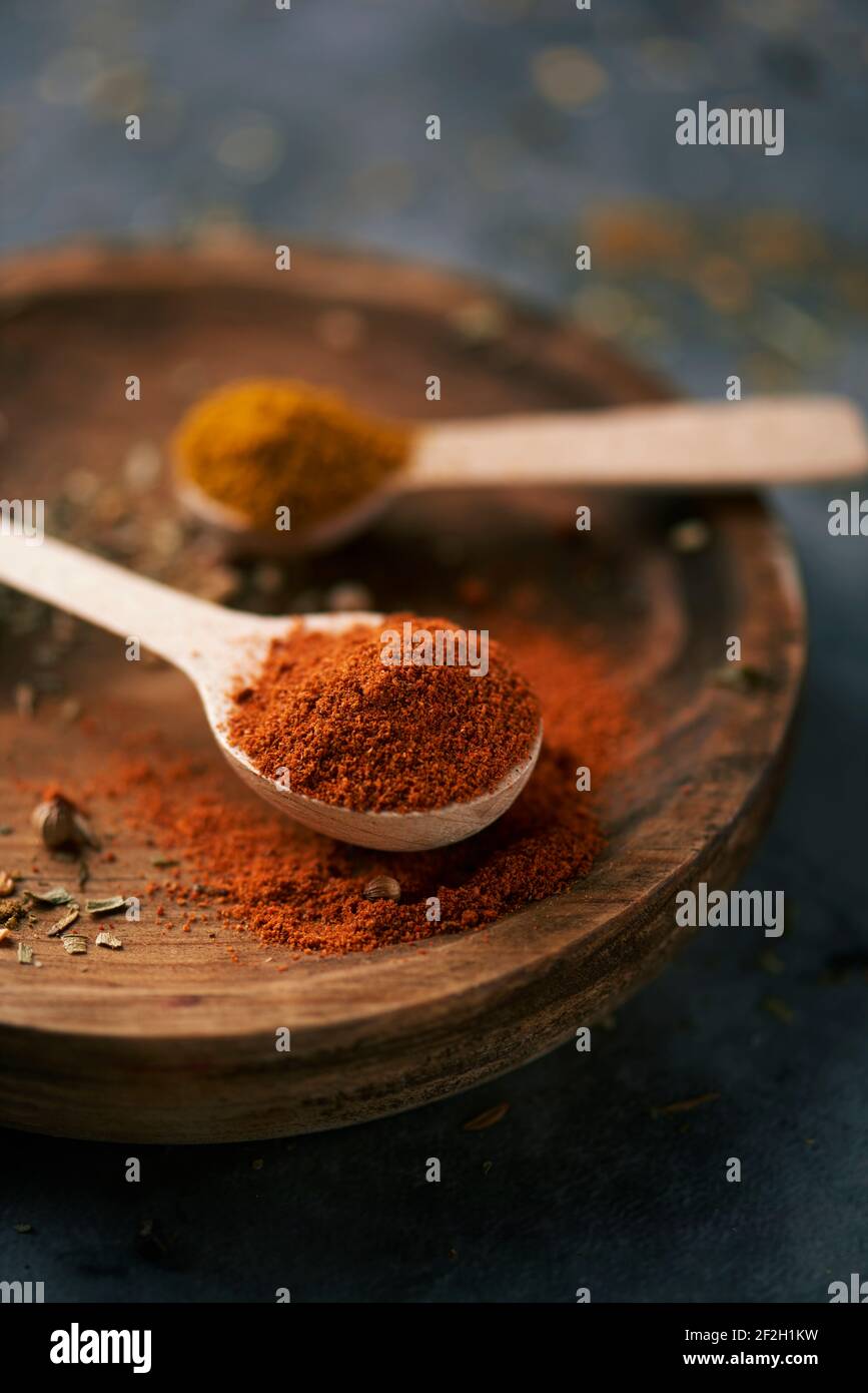 Nahaufnahme eines Holzlöffels voll rotem Currypulver neben einem anderen Holzlöffel voll regelmäßigem Currypulver, auf einem Holzteller, auf einem dunklen ston platziert Stockfoto