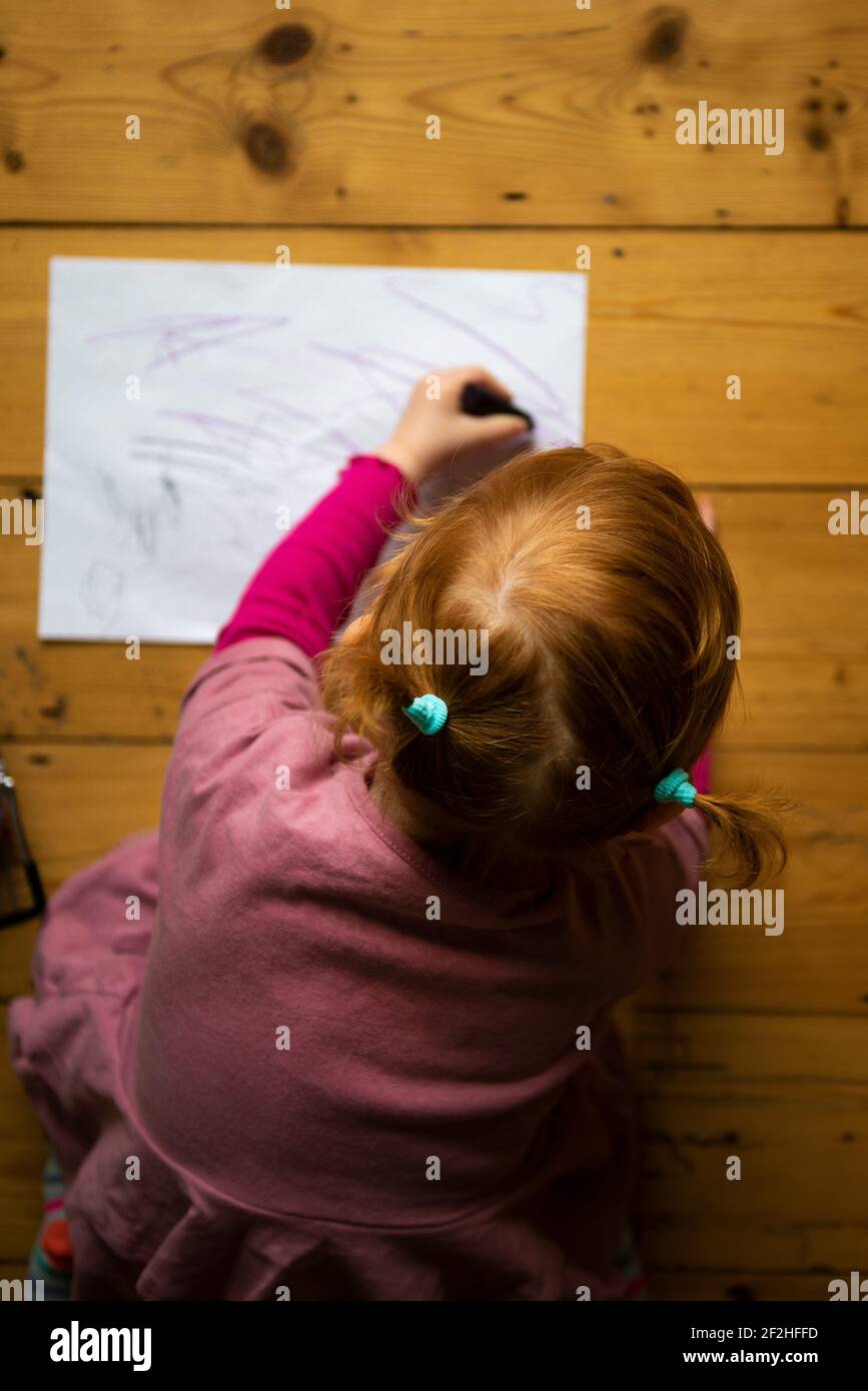 Ein kleines Mädchen in rosa gekleidet und mit ihren Haaren in Zöpfen sitzt auf einem Holzboden Zeichnung auf weißem Papier mit bunten Buntstiften. Stockfoto