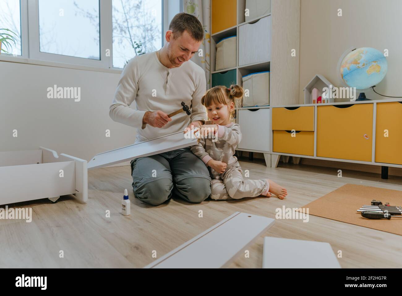 Kleines 4-jähriges Mädchen, das ihrem Vater hilft, das Kinderbett mit einem kleinen Hammer im Kinderzimmer zusammenzustellen. Stockfoto
