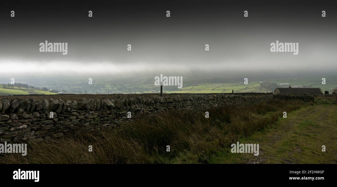 Eine unwirtliche Landschaft aus Trockensteinmauer, Bauernhaus und dunkelgrauem Himmel in den North Pennines, Weardale, County Durham, Großbritannien. Stockfoto