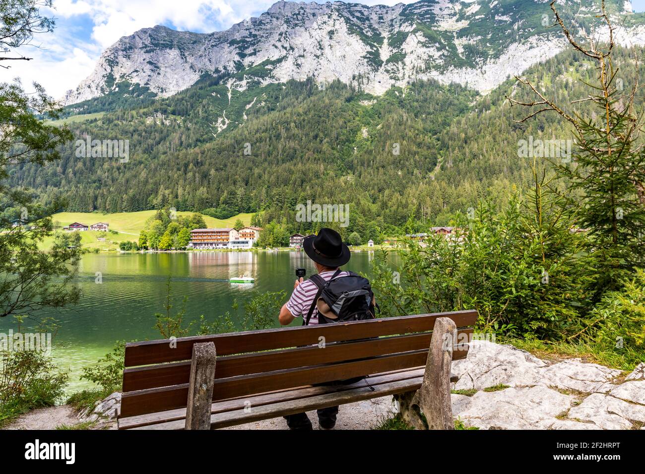 Wanderer sitzt auf einer Bank und filmt die Landschaft mit der kleinen Kamera, Hintersee, Ramsau, im Hintergrund Reiteralpe, Berchtesgaden, Berchtesgadener Alpen, Nationalpark Berchtesgaden, Berchtesgadener Land, Oberbayern, Bayern, Deutschland, Europa Stockfoto