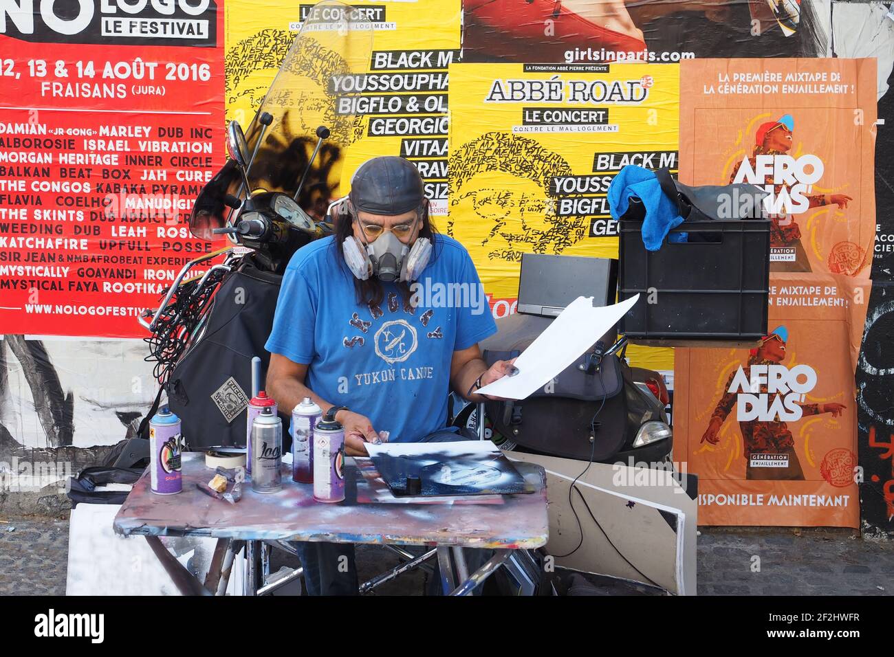 Bärtiger Straßenkünstler, der eine Atemschutzmaske an seinem Schreibtisch trägt, mit seinen Arbeiten, die mit Farben in Aerosoldosen gemacht wurden, mit Anzeigen von Musikkonzerten im Hintergrund Stockfoto