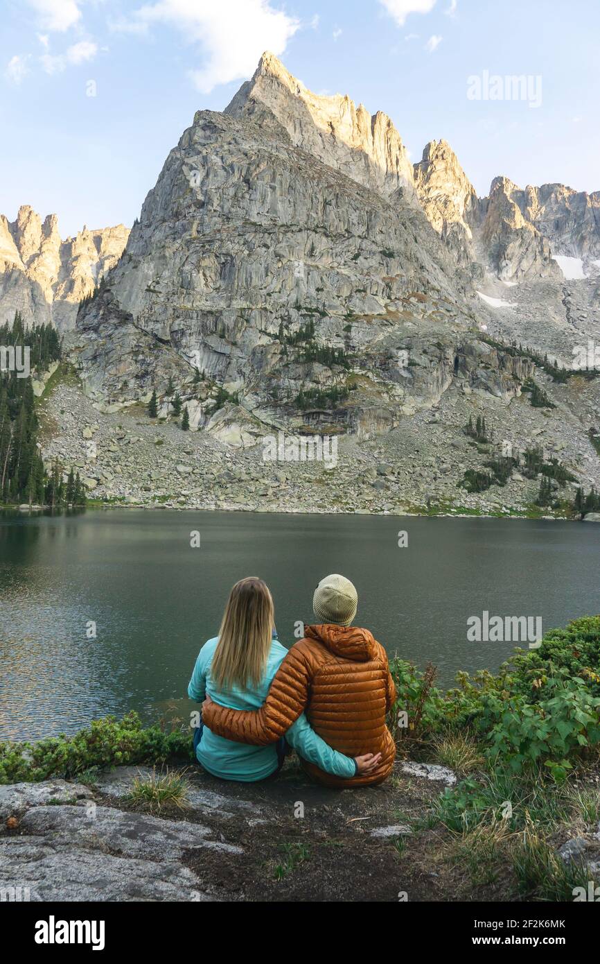 Rückansicht eines Paares, das während des Aufenthalts den Lone Eagle Peak ansah Mit den Armen am Seeufer sitzen Stockfoto