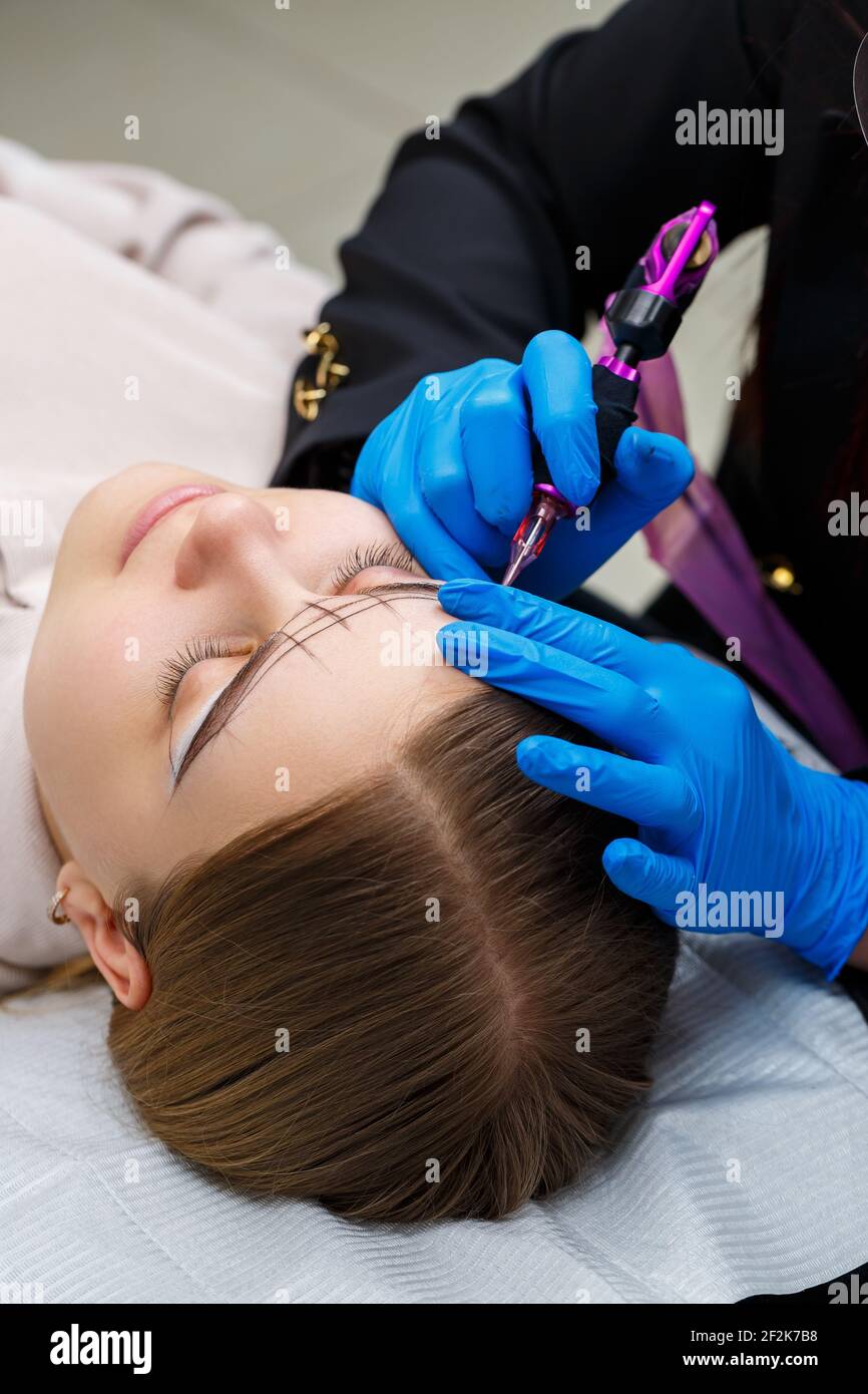 Kosmetikerin tätowiert die Augenbrauen einer Frau mit spezieller Ausrüstung während des permanenten Make-up, Nahaufnahme. Stockfoto