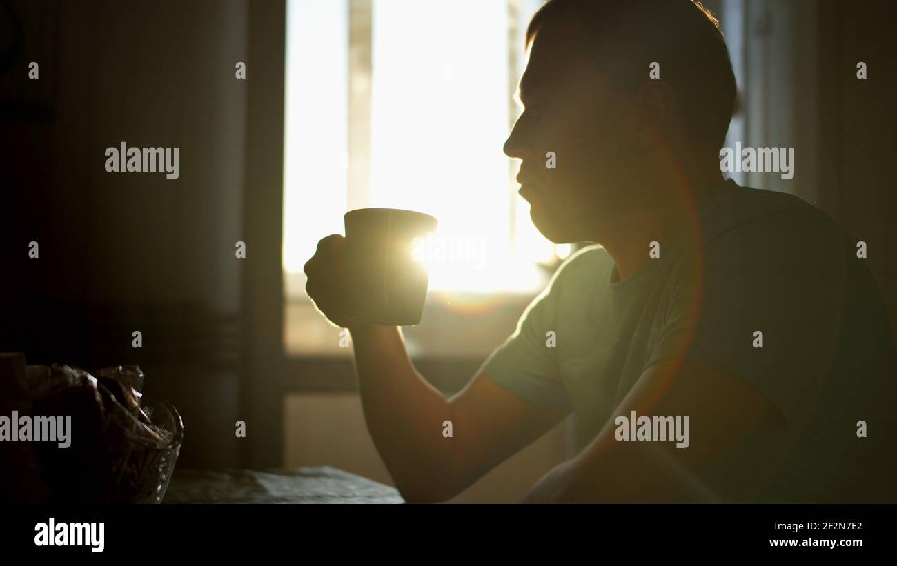 Silhouette eines jungen Mannes in den Sonnenstrahlen der Morgendämmerung trinkt heißen Kaffee, während zu Hause sitzen, Kamerabewegung Stockfoto