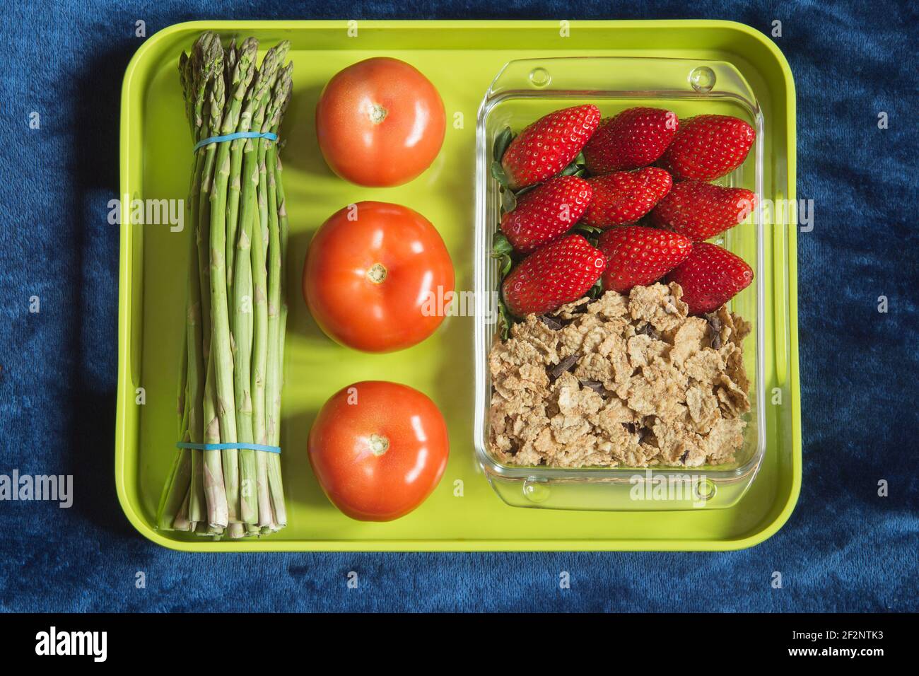 Eine Glasform gefüllt mit Erdbeeren, Vollkornprodukten und dunkler Schokolade auf einem Tablett mit Tomaten und wildem Spargel. Gesunde Ernährung. Stockfoto